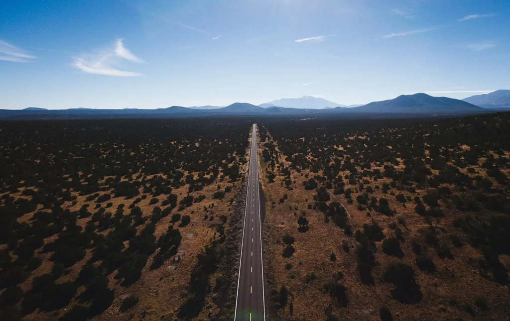 Carretera US-180 recta entre bosque bajo y montañas cerca de Flagstaff, ruta del norte con placa temporal en Arizona